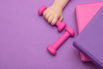 Sports equipment for fitness and yoga - dumbbell, blocks, one of which is held by a child's hand on a lavender-colored sports mat. Place for your text.