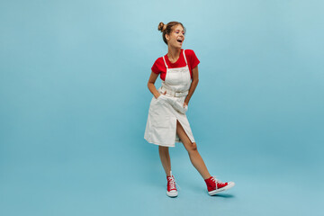 Full length cute young caucasian woman laughing on blue background while holding her hands in pockets. Pretty model with light-brown tufts on head, wearing red T-shirt and white sundress.