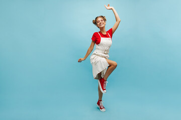 Full length energetic young caucasian girl posing over blue studio background. Slender blonde with two tufts of hair is wearing red T-shirt and white sundress. Lifestyle, female beauty concept