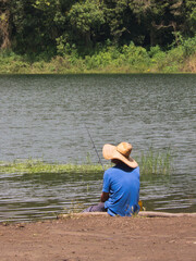 Hombre de camisa azul con sombrero sentado a la orilla de un lago pescando