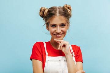 Close-up of stylish young caucasian woman looking at camera while holding her chin over blue background. Smiling girl with two bundles of hair, dressed in casual clothes.