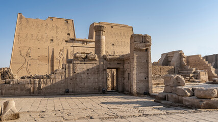 The Temple of the Horus in Edfu. Against the background of the blue sky, high walls with carvings, a column, ancient stones are visible. Egypt - obrazy, fototapety, plakaty