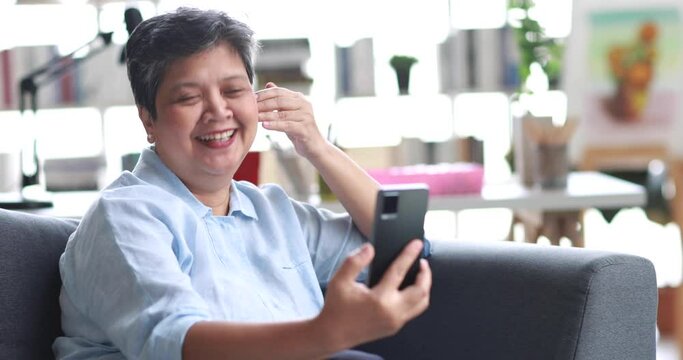 Happy mature female smiling and talking while sitting on couch and making video call via smartphone on weekend day at home