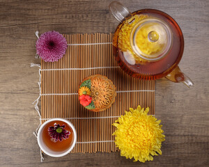 Colorful flower decorated moon cake Chinese mid autumn festival tea in small teacup glass teapot daisy chrysanthemum mum flower red yellow pink purple violet bamboo food mat on yellow background