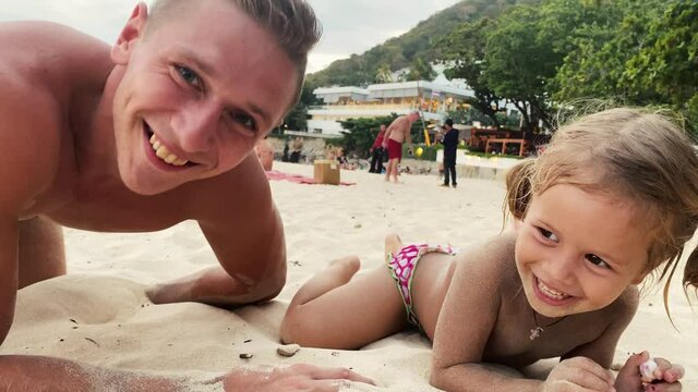 Dad and daughter run along a sandy beach at sunset. They fall on the sand. Dad makes face. The little daughter laughs and climbs on Dad's back. She smiles. Happy childhood. Family. Love