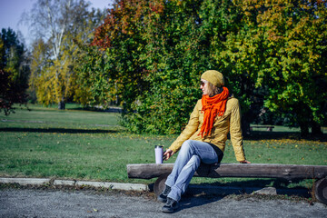 Pretty european woman in yellow jacket and red scarf sitting on wooden bench in deserted park. Enjoying solitude and sunny autumn weather. Blurred plant background, copy space.