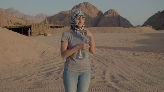 Woman in checkered keffiyeh drinking water of plastic bottle standing in the desert. Caucasian female tourist quenching thirst during desert adventure.