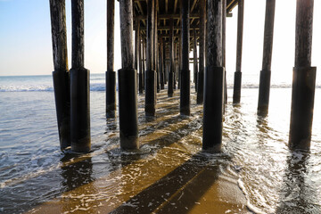 a shot of the legs underneath a brown wooden pier at the beach with waves rolling in from the vast...