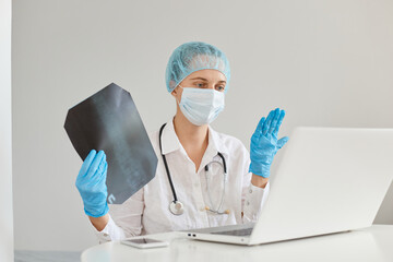 Indoor shot of female doctor with x-ray in hands , working at the office desk in front of laptop, having online colloquium, consults with colleagues about the treatment of pneumonia of patient.