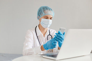 Indoor shot of concentrated woman doctor therapist wearing gown, surgical mask, medical cap and gloves, working in front of computer, holding smart phone in hands, dialing number.