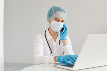 Portrait of serious woman doctor wearing gown, surgical mask, medical cap and gloves, posing in front of notebook, talking phone with college, working in hospital.