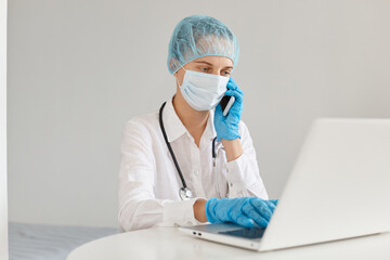 Horizontal shot of woman doctor therapist wearing gown, surgical mask, medical cap and gloves, working in front of notebook and talking via mobile phone.