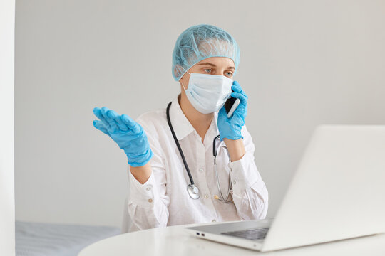 Confused Helpless Woman Doctor Wearing Gown, Surgical Mask, Medical Cap And Gloves, Posing In Front Of Notebook, At Desk, Talking Phone, Spreading Hand Aside.