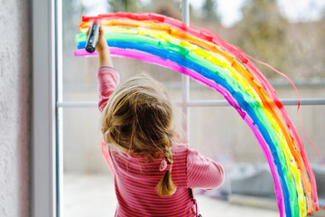 Adoralbe little toddler girl with rainbow painted with colorful window color during pandemic coronavirus quarantine. Child painting rainbows around the world with the words Let's all be well.