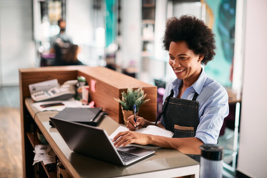 Happy African American hairdresser writes notes while working on laptop at her salon.