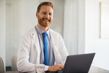 Happy doctor works on laptop at his office and looks at camera.