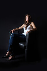 Full length portrait of a beautiful brunette woman sitting on black armchair in front of dark background