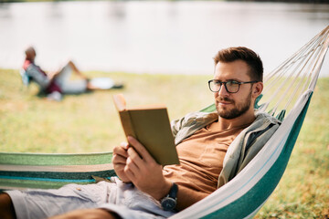 Fototapeta na wymiar Mid adult man reads book while relaxing in hammock in nature.