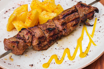 Grilled pork skewer with mustard sauce and crispy potato slices on white plate.