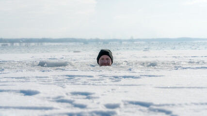 Ein Mann guckt aus einem Eisloch beim Eisbaden in einem zugefrorenen See bei Schneefall