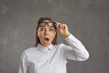 Amazed woman raises her glasses looking into the camera with a distrustful expression. Surprised and embarrassed woman with wide open mouth and eyes stands on a gray background. Banner.