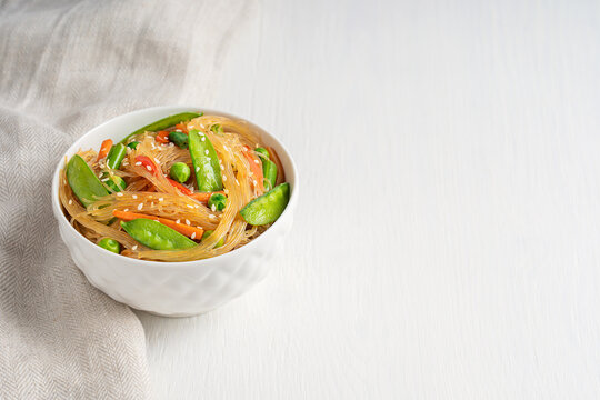 Japchae savory vegetarian korean popular dish of stir-fried glass noodles and vegetables such as green peas, bean and carrot served in bowl with textile on white wooden table. Image with copy space