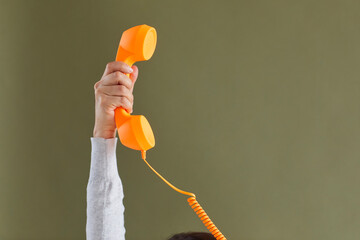 Human hand holding bright vivid orange landline phone receiver. Cropped shot of young lady raising up retro telephone receiver against khaki green blank copy space background. Customer support concept - obrazy, fototapety, plakaty