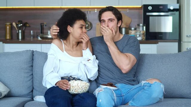 Skocked couple of caucasian man and african american young woman watching horror movie film at stylish apartments, sitting on sofa together with with popcorn at home