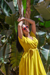 A Latina woman standing with her arms raised in a yellow dress looking up on a background of green plants