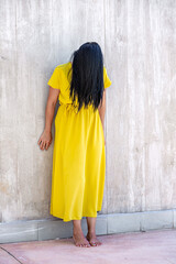 Fototapeta premium Adult woman with long black hair barefoot and standing leaning on a wall with her head tilted forward and her hair covering her face