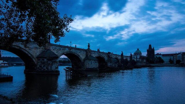 Blue morning timelapse of the Charles Bridge in Prague, Czech Republic next to the Vltava river with panoramic view of the river shore