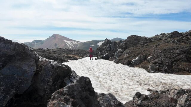Reveal of a hiker walking with a backpack on the patch of snow between black magma rocks in Landmannalaugar in Iceland