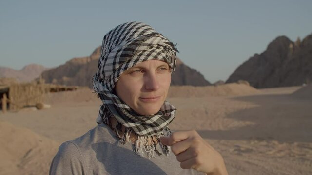 Close up of woman in checkered keffiyeh drinking water of plastic bottle standing in the desert. Caucasian female tourist quenching thirst during desert adventure.