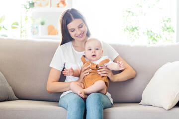 Portrait of attractive kind cheerful girl holding playing baby sitting on divan spending free time at light home flat house indoors