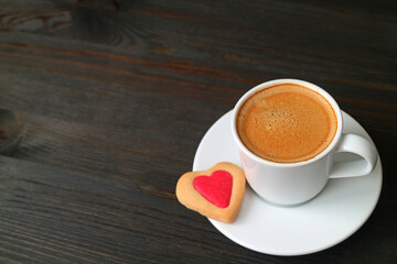 Cup of hot coffee with a heart shaped cookie on black wooden backdrop