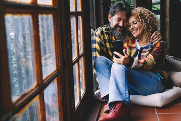 Fototapeta na wymiar Happy adult couple enjoy mobile phone call at home sitting on the floor and hugging with love and romance. Cheerful man and woman in cellular calling together