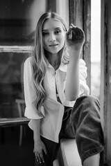 a beautiful girl in a white shirt with blonde hair is sitting on the windowsill of a large window