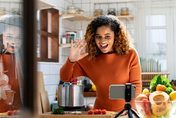Young African American woman looking at mobile phone and say hi on social media in kitchen at home,...