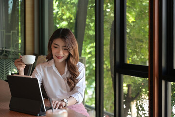 Photo of a young businesswoman drinking a coffee while sitting in front of a digital tablet at the meeting table.