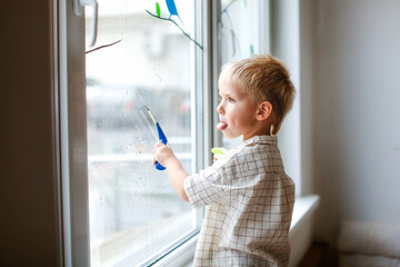 Fototapeta na wymiar Cute boy toddler blond toddler at the window washes the glass. Helping children with household chores and household chores, children's independence, Montessori classes for small