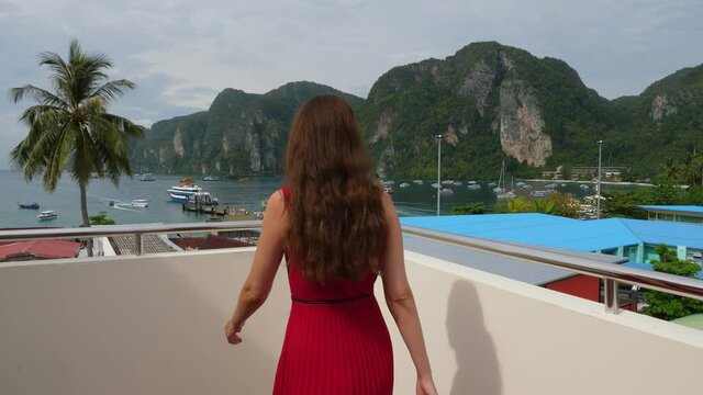 Woman walk at terrace to railings, stand and look to Tonsay bay and village buildings from height, moving camera follow behind and then move forth. Small town at Koh Phi Phi Don
