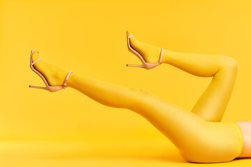 Long slim legs of young woman in yellow tights and stylish high heels shoes on yellow background. - obrazy, fototapety, plakaty