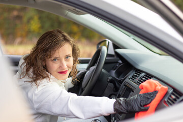 A beautiful young woman driver in a white jacket and black gloves wipes the dust in the car interior with a red rag on a bright warm autumn day. Selective focus. Portrait