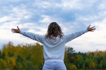 A woman in a white sweater with a hood stands in a field against the background of a forest and a blue sky with her arms outstretched wide on a warm autumn day. Selective focus. Close-up. Portrait