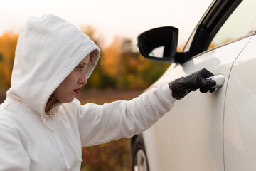 A beautiful young woman in a white sweater with a hood and black gloves opens the car door to steal it. Selective focus. Close-up. Portrait