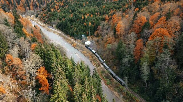 Aerial drone view of the moving steam train Mocanita in a valley along a river, hills covered with yellowing forest, Romania