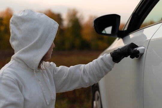 A beautiful young woman in a white sweater with a hood and black gloves opens the car door to steal it. Selective focus. Close-up. Portrait