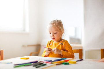 Cute sunny girl child blonde with a ponytail at the table draws with multicolored felt-tip pens,...