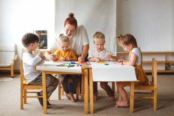 cheerful teacher woman with a group of cute preschool children at the table draw on paper with multicolored felt-tip pens, montessori and creativity, creative development and lesson in kindergarten