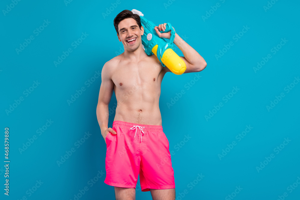 Wall mural Photo of funny adorable young man naked torso holding water gun smiling isolated blue color background